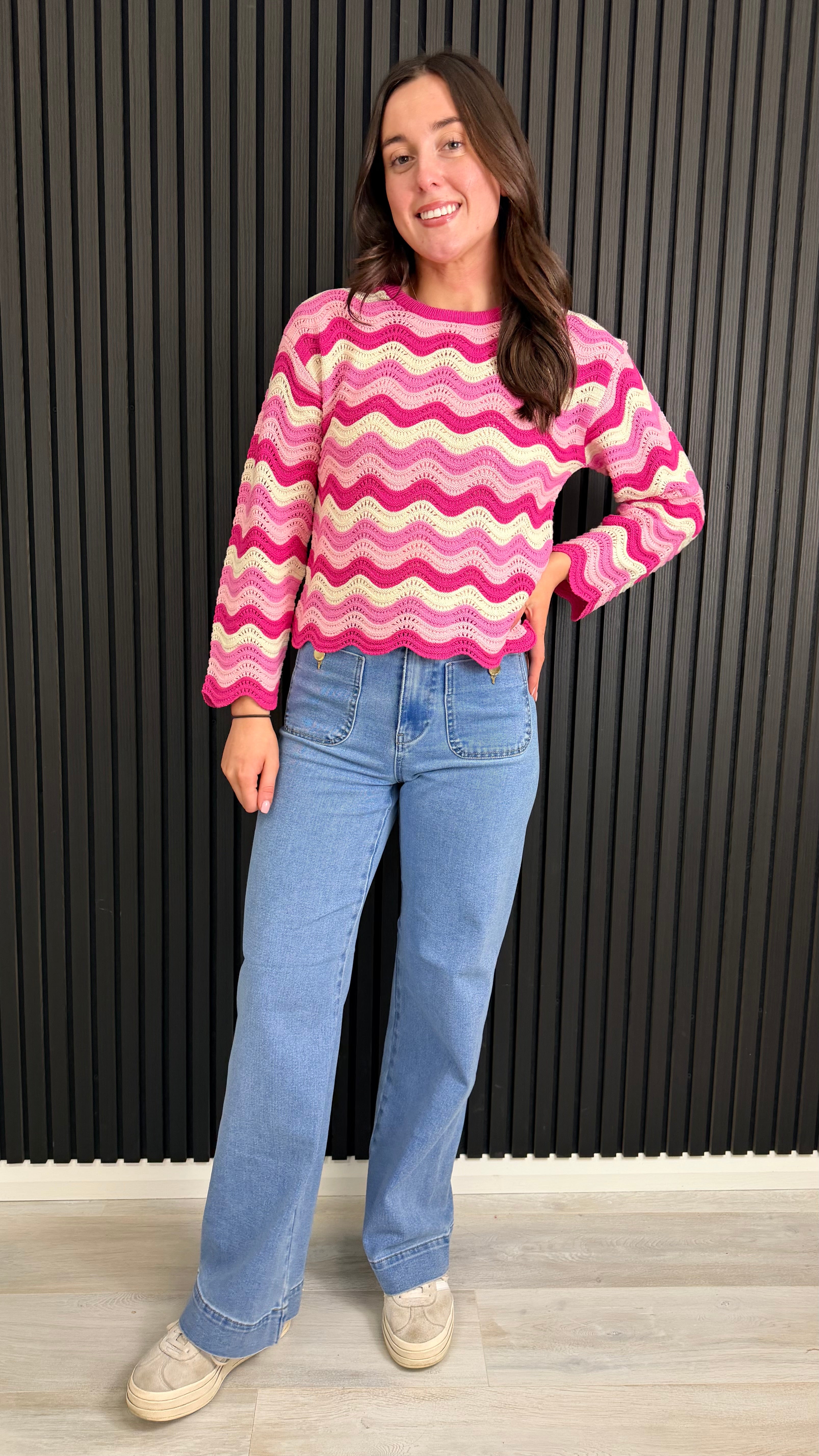 Full length woman wearing a pink and cream zigzag knit jumper styled with blue jeans.