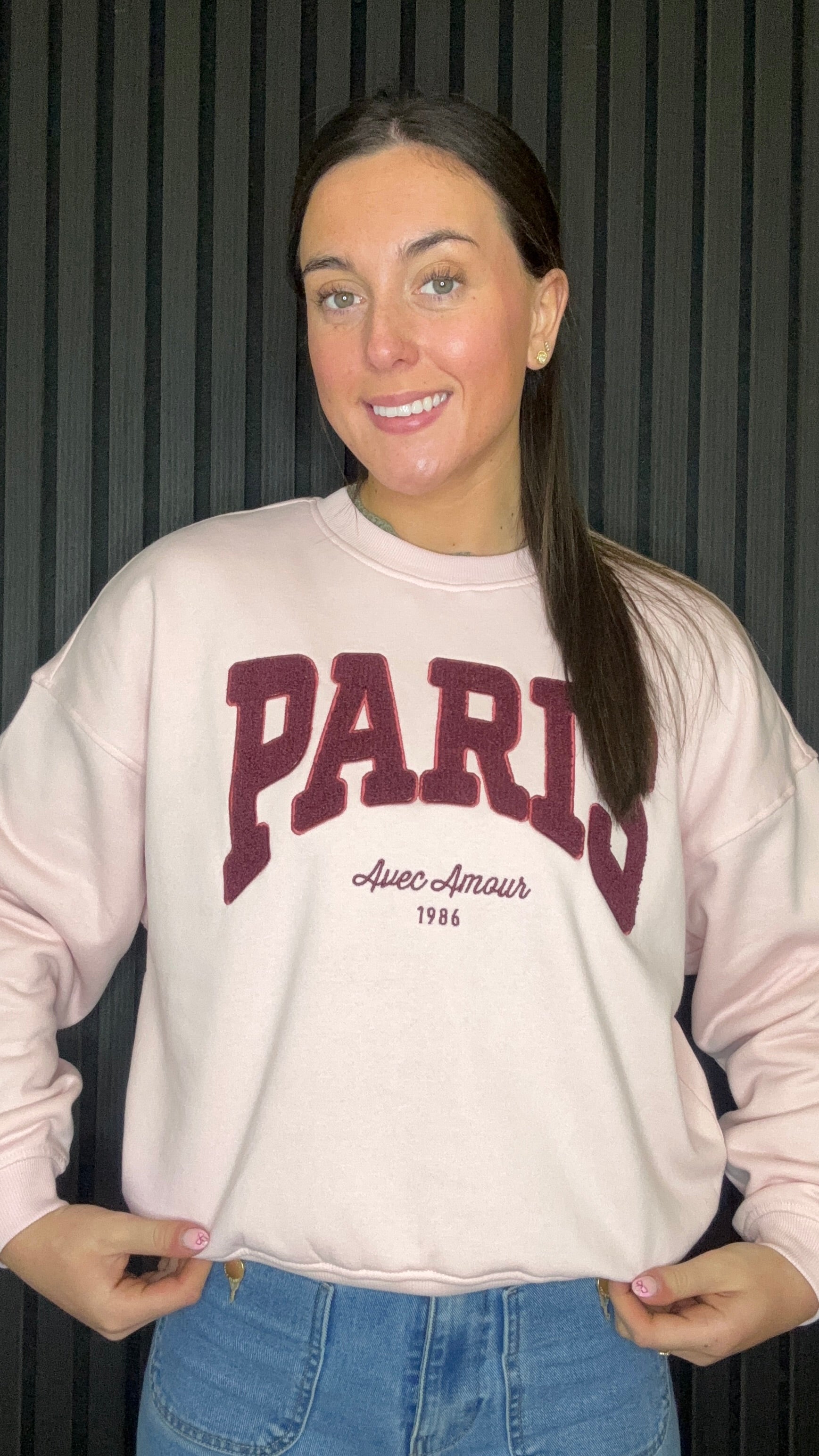 Model wearing chalk pink Paris slogan sweatshirt with burgundy lettering, crew neck and relaxed fit, paired with blue jeans.
