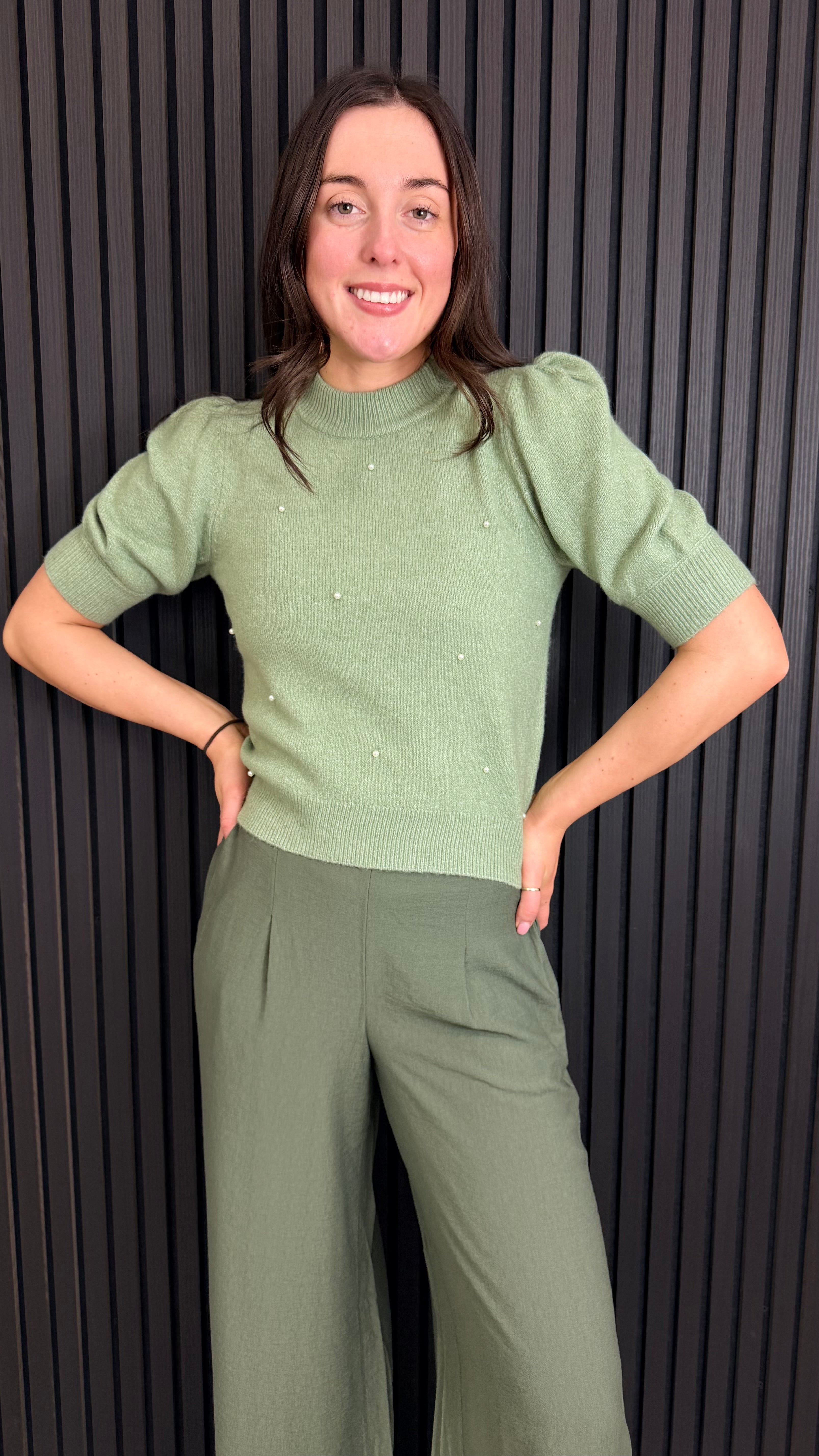 Full length Woman wearing a sage green short-sleeve knit top with pearl detailing.