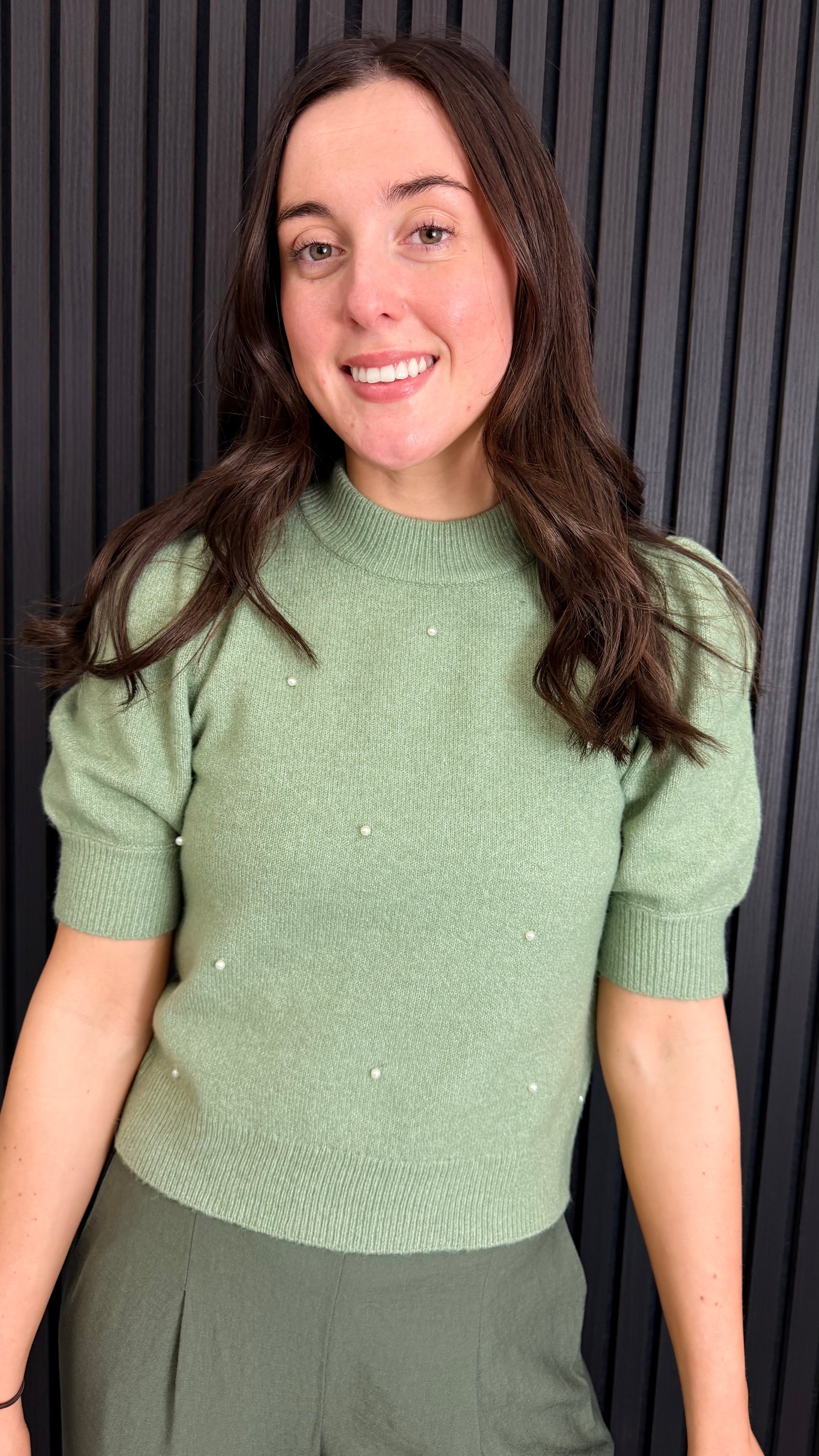 Woman wearing a sage green short-sleeve knit top with pearl detailing.
